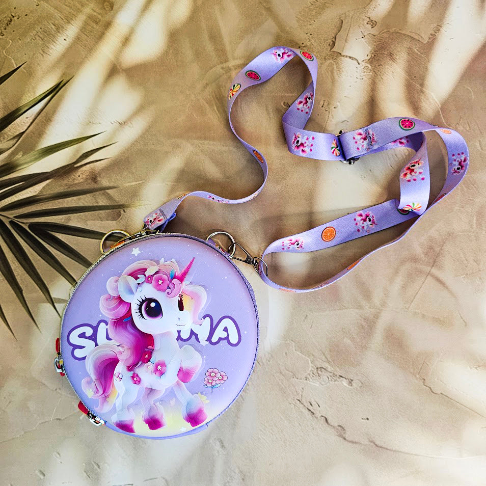 Two colorful, round kids’ sling pouches with attached lanyards are placed on a sandy beige background with a tropical leaf shadow. The left pouch is purple and features an adorable cartoon unicorn with a pink mane, large sparkling eyes, and the name "SHYAMA" written in white. It has a lilac lanyard decorated with candy prints. The right pouch is bright blue with a fierce green cartoon T-Rex dinosaur illustration labeled “DINOSAURS” in bold yellow text. Its lanyard is blue with black dinosaur silhouettes. Bo